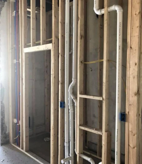 Exposed wooden wall studs with white PVC plumbing and electrical wiring during construction.