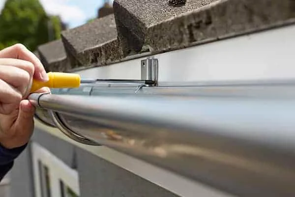 Person using a screwdriver to install or adjust a metal gutter on a roof with dark tiles.